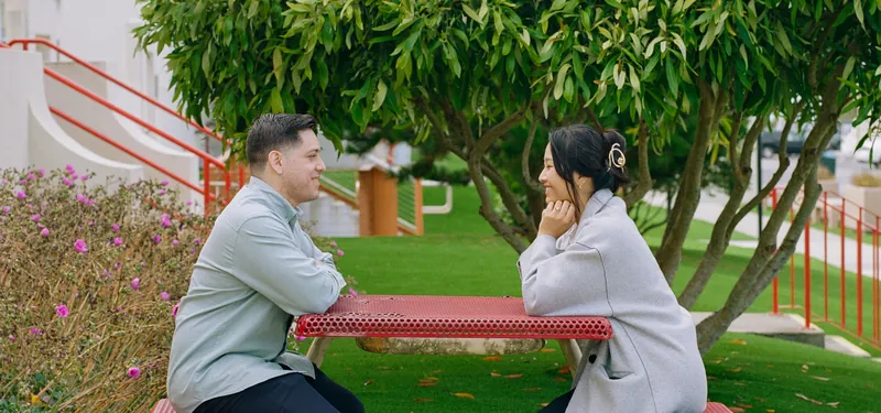 Eden and Luis sitting across from each other at a red picnic table under a tree