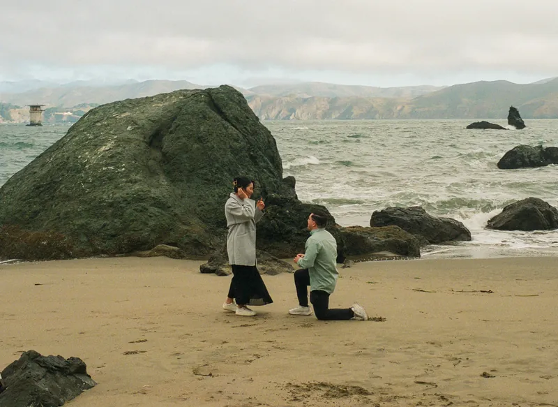 Luis on one knee proposing to Eden on the beach beside a large rock
